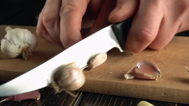Close-up of hands slicing garlic cloves on a wooden cutting board with additional garlic pieces scattered around in a kitchen setting