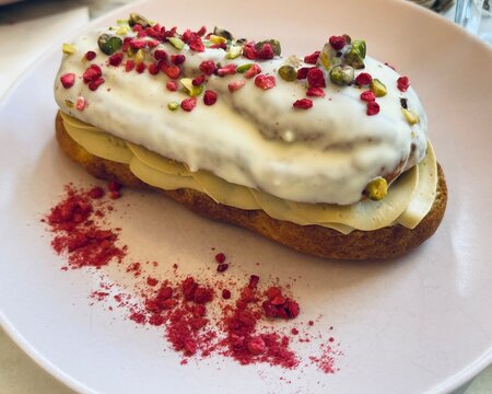 Gluten free and lactose free &eacute;clair dessert with white chocolate glaze, pistachios and dried raspberry pieces, served on elegant plate in cafe setting.