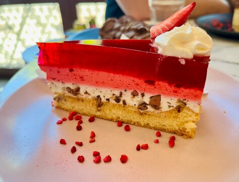 Layered strawberry jelly cake with creamy filling and sponge base, decorated with whipped cream and fresh strawberry slice. Colorful dessert served on white plate in cafe setting, close up.Gluten-free