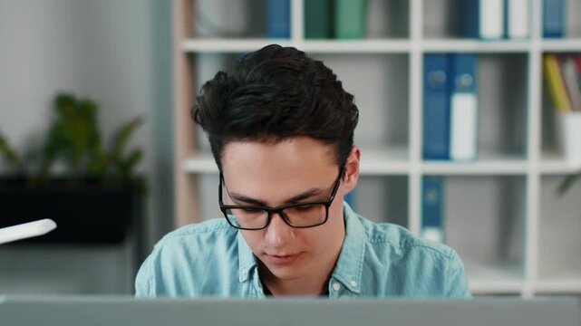 A young man with light skin and glasses is focused on his computer screen in a contemporary office environment. Shelves in the background hold various folders and plants.