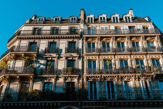 Classic Paris residential building with wrought iron balconies, tall windows and mansard roof illuminated by sunlight against clear blue sky