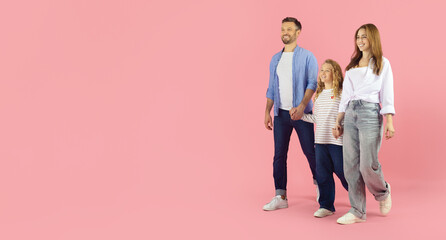 Naklejka premium Happy caucasian family of three walking together on pink studio background with copy space. Father, mother and daughter stroll hand in hand in casual outfits looking away