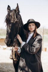 Obraz premium Woman stands with black horse wearing a hat and a suit in a rural area during daylight hours