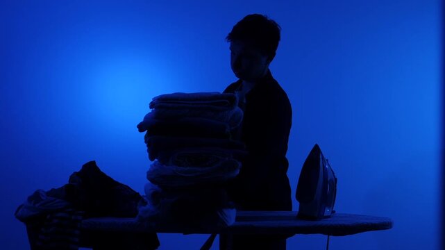 Man suffering from a headache and feeling stressed by household chores, standing behind a large pile of clothes to iron on an ironing board in a dark blue environment
