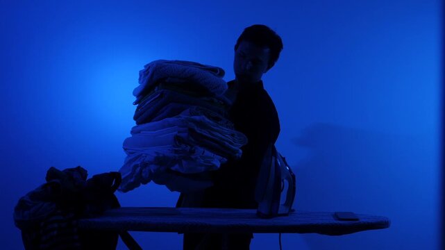 Man in silhouette carrying a large pile of fresh laundry, standing next to an ironing board with an iron, performing household chores in a domestic setting under blue light