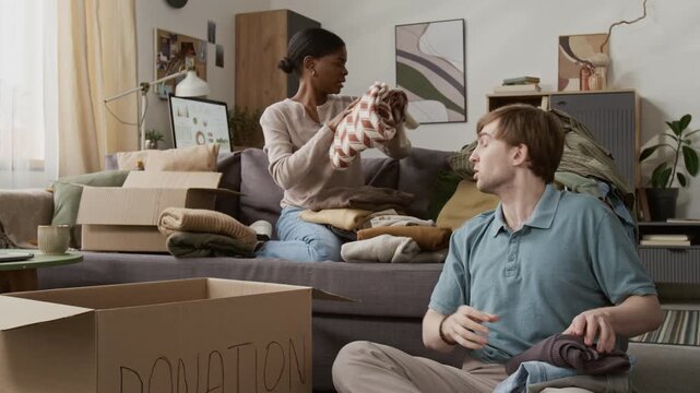 Multiethnic young couple sorting clothes and discussing them while deciding which items to donate, man putting them into cardboard box, girl sitting on couch in background