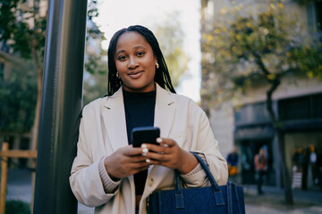 Fototapeta premium Confident woman professional crossing a city street with a smartphone and bag, successful female entrepreneur on the move in an urban setting, nomadic lifestyle and mobility concept.