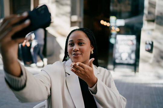 Radiant woman professional blowing a kiss while taking a selfie at a city cafe, happy lady freelancer recording content for social media vlogs, modern mobility and startup work culture.