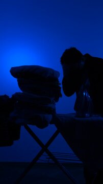 Vertical video man silhouette ironing clothes at board against deep blue backdrop, portraying fatigue and daily household drudgery