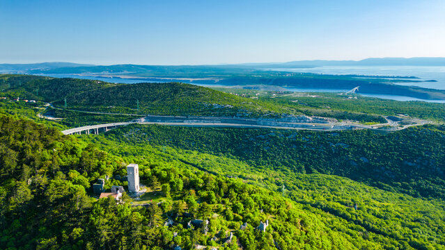 Aerial view of lush green hills, historic Trsat tower, winding roads, and the iconic Krk Bridge spanning the clear blue Kvarner Gulf in Croatia. Spring beauty. Highway A7
