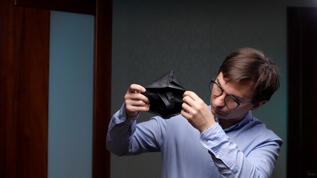 Young man in glasses and a light blue shirt shows an empty wallet, expressing financial hardship, debt worries, unemployment and the stress of running out of money