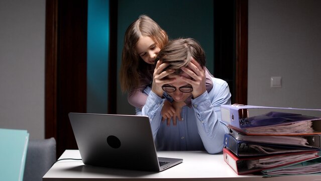 Overwhelmed father working late on laptop in home office, burned out and stressed as his young daughter hugs him, seeking attention and comforting amid demanding workload