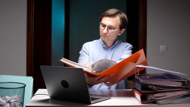 Business man wearing glasses checking financial documents in an orange binder at his office desk with a laptop and a stack of folders, working late in the evening
