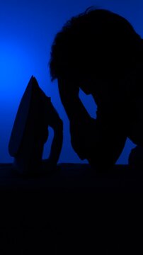 Vertical video man silhouetted ironing with visible fatigue and reluctance against a vibrant blue backdrop, conveying household drudgery