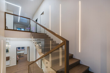Stairs to the second floor in the lobby of a luxurious mansion. White wall decor,  parquet, dark wood staircase with glass railings. © Alhim