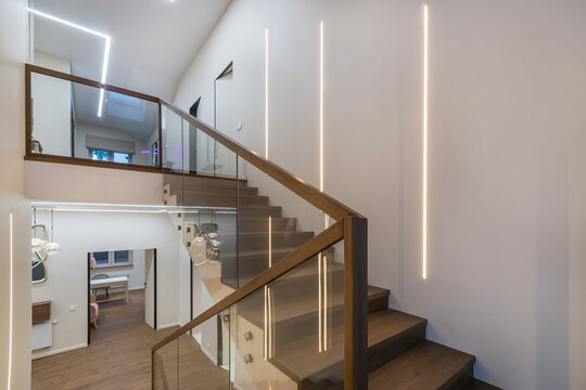 Stairs to the second floor in the lobby of a luxurious mansion. White wall decor, parquet, dark wood staircase with glass railings.