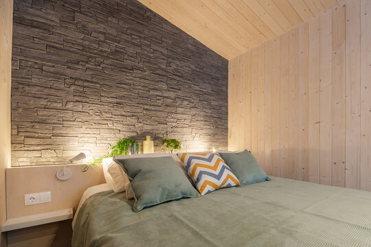 Stylish bedroom interior in eco-style. Wooden walls, imitation stone. A wide bed with a bedspread and pillows.
