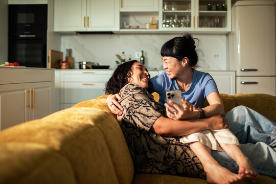 Two women relaxing with smartphone on cozy sofa in modern home kitchen
