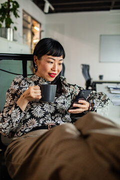 Businesswoman drinking coffee and using smartphone in modern office