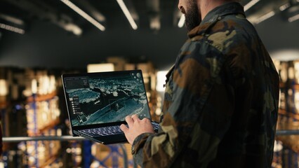 Male army soldier ensuring surveillance in secure military warehouse, personnel monitoring drone dashboard on laptop. Defense logistics to ensure supply chain for military base operations. © DC Studio