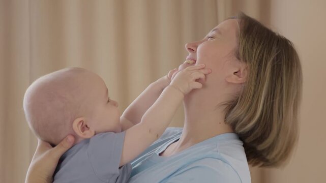 Caucasian mother laughs as baby touches face, playful fingers on cheeks, mutual smiles, light curtain backdrop, tender interaction, bonding through touch, joyful