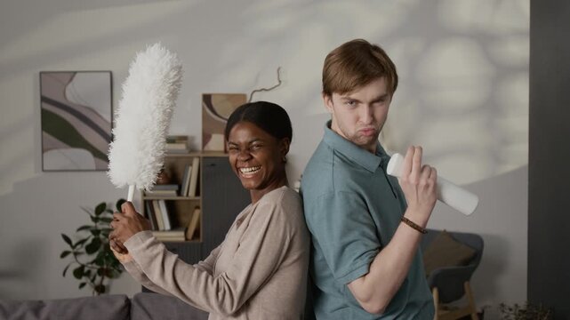 Portrait of smiling Black girl with feather duster and Caucasian man holding white sprayer posing for camera pretending to be ready to fight in living room