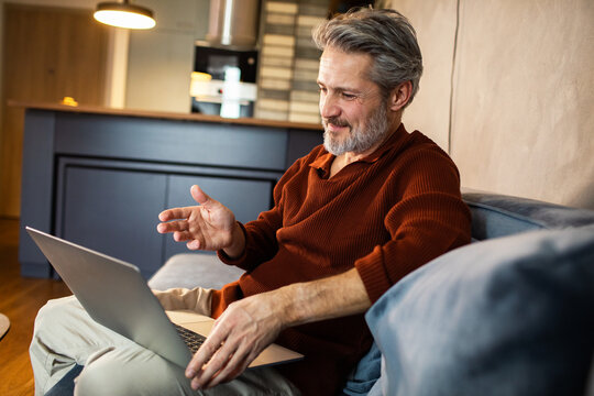Mature man video calling on laptop from home couch