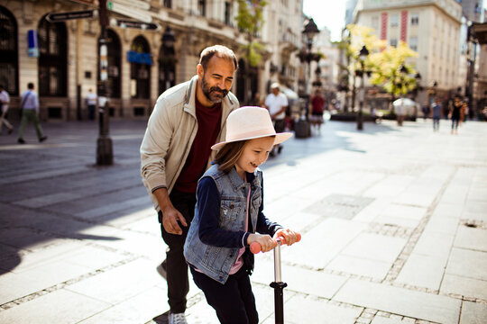 Father teaching daughter to ride scooter on sunny city street