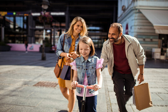 Parents watching daughter ride scooter on city street