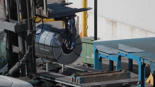 Circle of metal roll is lifted by a crane at a loading dock during daytime in a busy industrial area