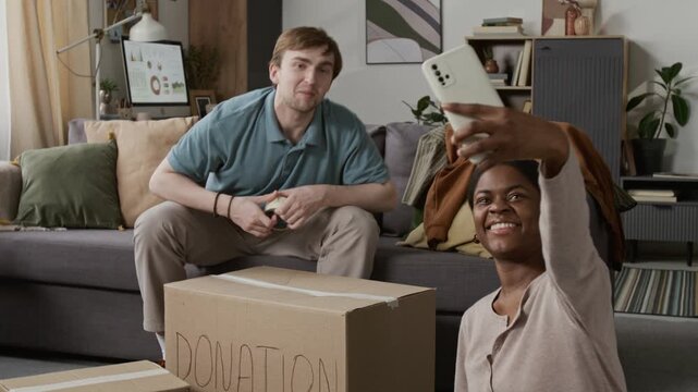 Positive Black girl holding smartphone taking selfie picture with smiling Caucasian boyfriend after he sealing large donation box in living room