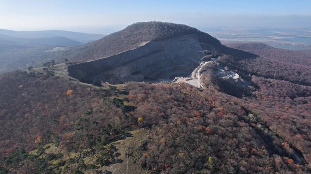 Flight over of a large open pit quarry with terraced excavation roads surrounded by forested hills in a rugged autumn landscape, Slovakia.