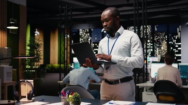 African american man, walking with laptop, office professional evaluating operations. Finishing tasks with notebook, Business manager employee working late at night to resolve complaints.