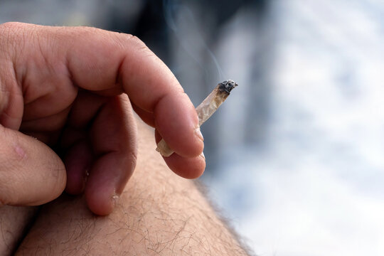 Close-up of a person's hand holding a cannabis joint, emitting smoke. Representing addiction, drug use, or relaxation