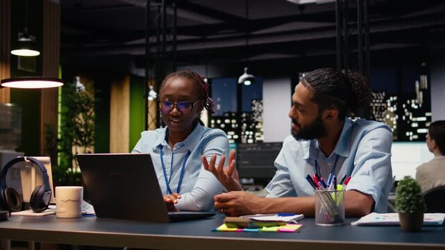 Diverse team of businesspeople at desk using laptop evaluating metrics, teamworking concept late at night. Colleagues working late at night on notebook to assist online clients, finishing tasks.