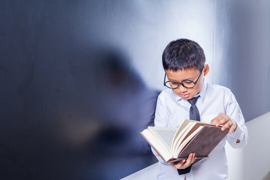 Smart Schoolboy with Glasses Concentrating and Reading a Book by Chalkboard