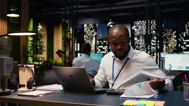 African american manager analyzes company data for accounting task, reviewing administrative business intel and building action plan. Employee at desk working late, project management.