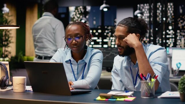 Diverse manager and assistant reading inbox emails on laptop late at night. Team analyzing data on notebook and interpreting statistical information, ensuring accounting assignments.