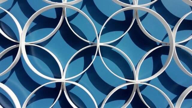 A minimalist architectural detail features white interlocking circles mounted against a solid blue surface. The bright sunlight casts sharp, curved shadows, creating a strong sense of depth and geomet
