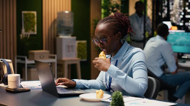 African American woman struggling with compulsive overeating, working on laptop. Employee with unhealthy eating habits serving junk food with carbs, reflecting mental health and burnout.