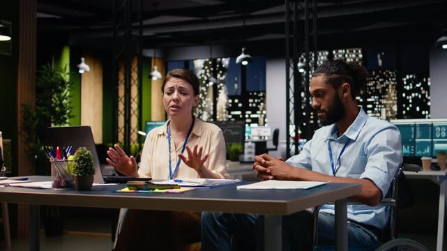 Corporate diverse coworkers team, wheelchair user and woman, collaborating late at night on a business assignment. Developing action plan, emphasizing inclusion and effective project management.