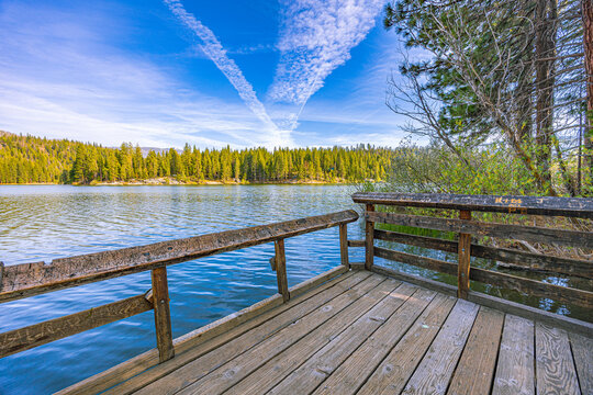 A wooden viewing deck extends over the calm blue waters of Hume Lake, surrounded by dense pine forest under a bright blue sky with contrails.