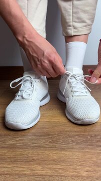 Close up of a woman tying shoelaces on white sneakers indoors. Sport shoes on wooden floor. Preparing for workout, fitness, jogging, training or daily casual activity.