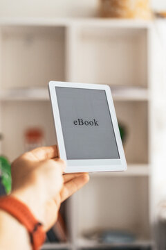 Close up of hand holding electronic book reader near shelves at home. Minimal lifestyle scene showing convenient reading, education access and modern technology for students and readers.