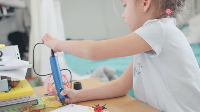 Little girl is using a 3D pen on a table filled with craft supplies. Room is bright and child is focused on creating. Tools and drawings are nearby.