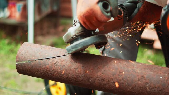 Industrial employee uses grinder to cut metal pipe at outdoor worksite. Scene captures technical precision, safety equipment and maintenance work for industrial systems. Grinding sparks