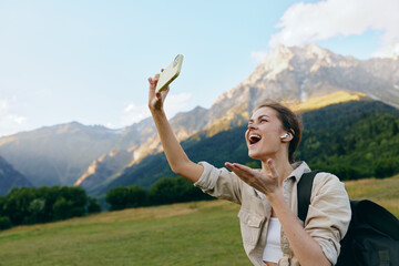 Fototapeta premium Cheerful woman outdoors, smiling while taking a selfie in a sunlit field, mountains in the background, showcasing a relaxed lifestyle and natural colors