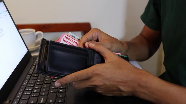 Unrecognizable Indonesian man checking cash money from his wallet at a work desk.
