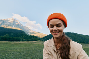 Fototapeta premium A smiling woman in a beige jacket and orange beanie enjoys a sunny outdoor moment in a green meadow with distant mountains, capturing a relaxed, casual lifestyle.