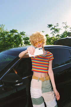 Young woman with curly blond hair takes a sunny selfie beside a dark car, wearing a striped crop top and patchwork pants, radiating carefree confidence in a casual outdoor setting.